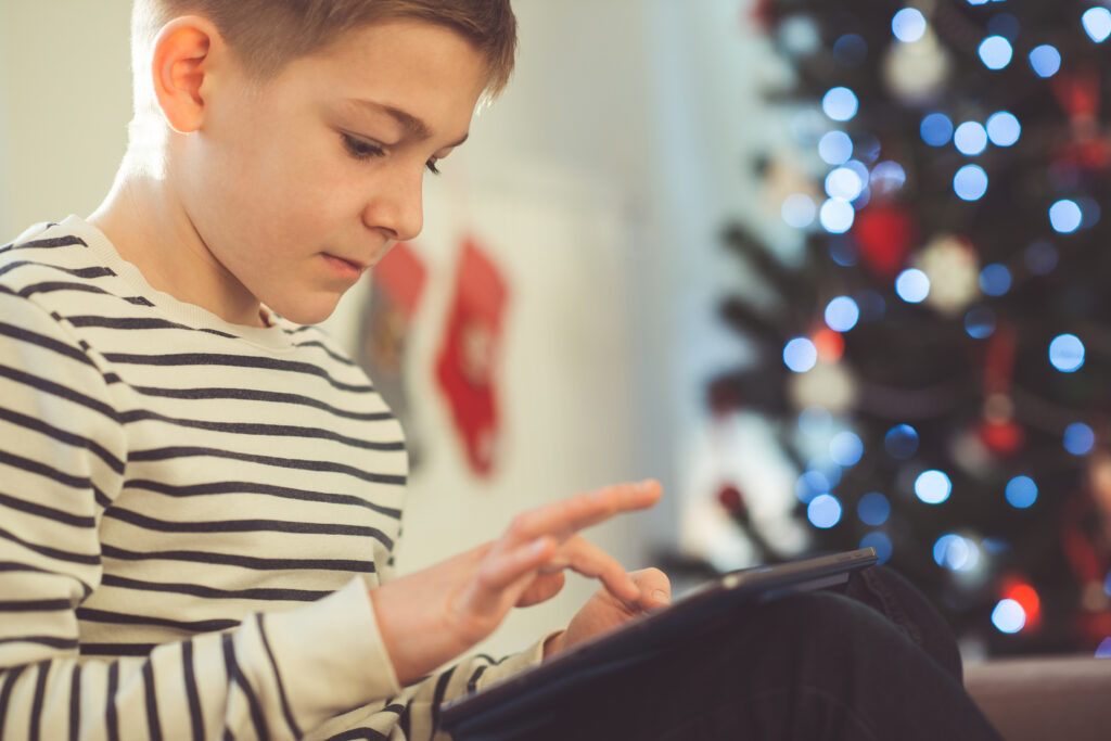 Teenager boy playing or studing with laptop near decorated christmas tree in living room at home