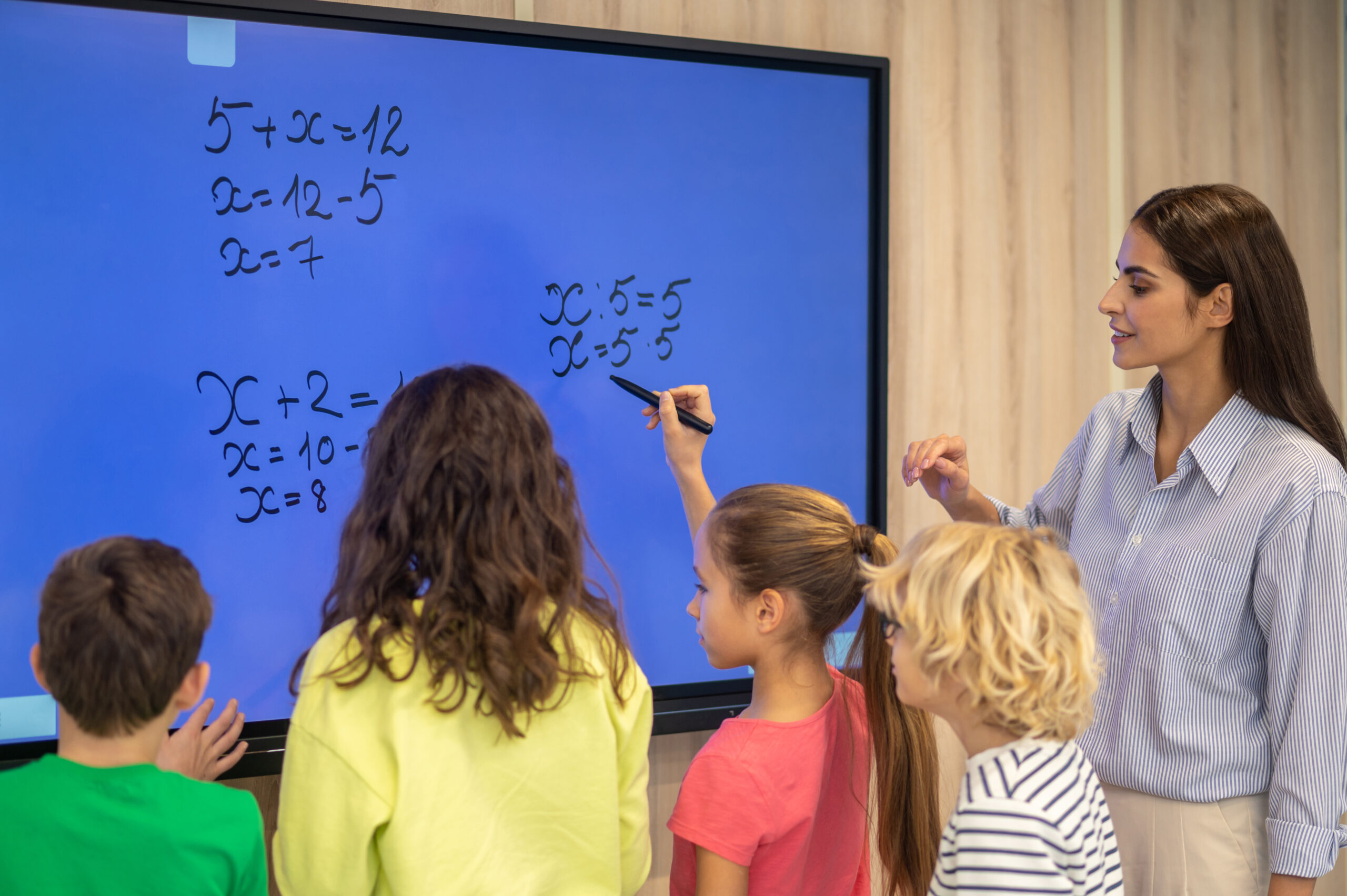 Equation solution. Boys and girls standing with backs to camera writing on blackboard and attentive teacher watching at math lesson